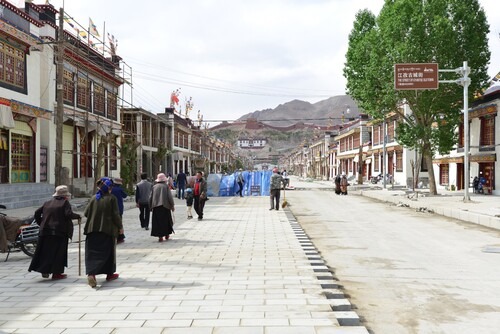 Prayer flags and PRC flags flutter above Gyantse Old Town, pictured during on-going redevelopment for tourism. Pelkhor Chödé Monastery looms in the background. People are pictured walking down the patchwork street and construction workers and their equipment can be seen. Faces are blurred.